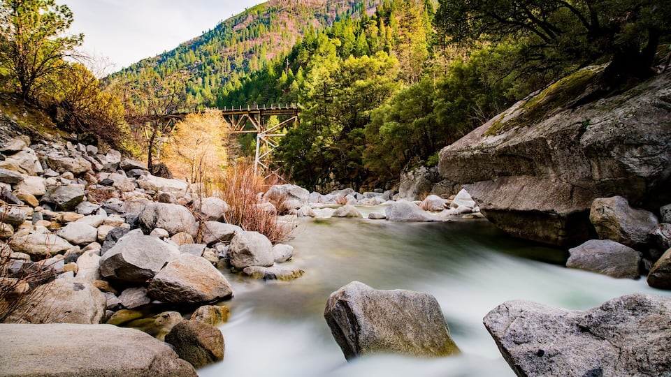 A river flowing through a rocky landscape with a wooden bridge and a forested hill in the background.