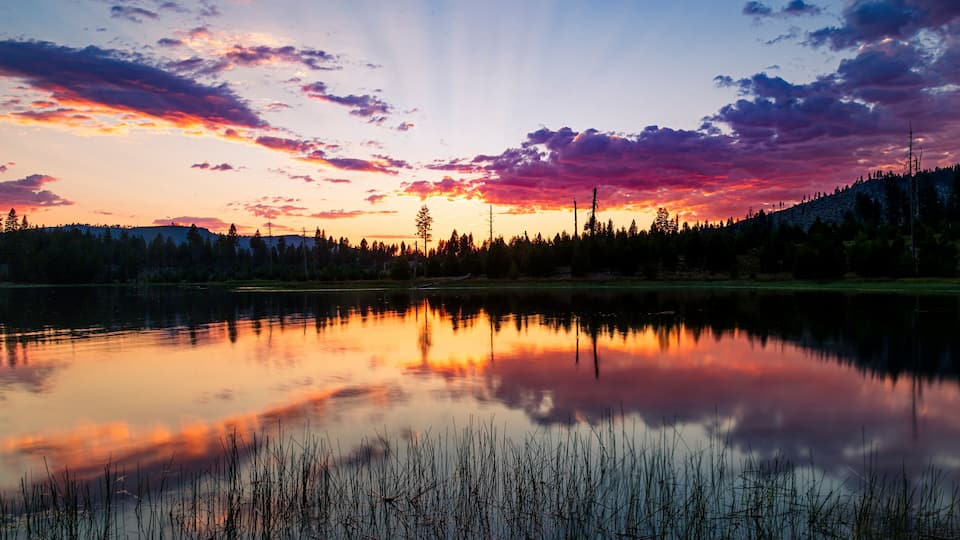 A calm lake reflecting a sunset with a forested mountain in the background.