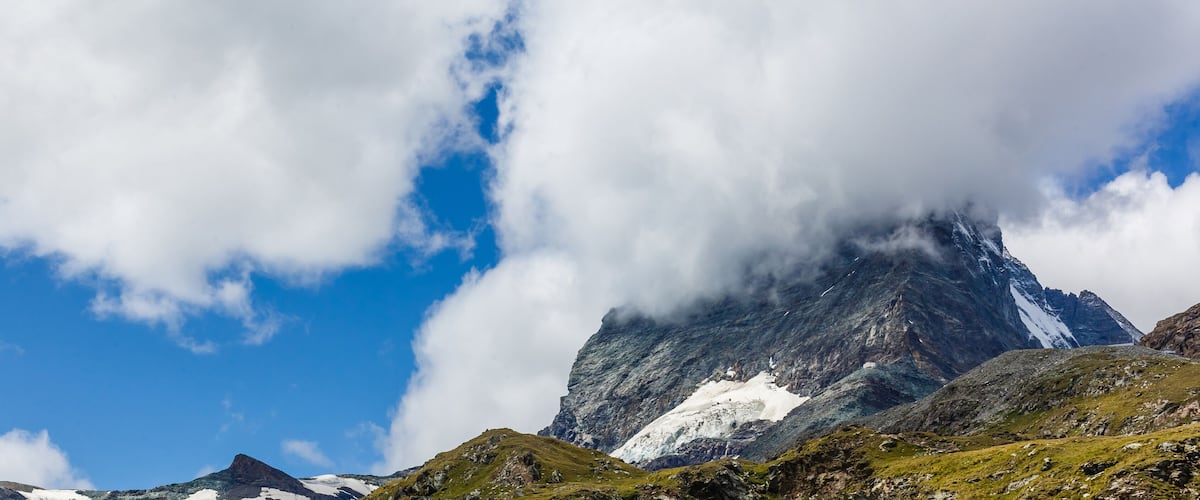 A mountain landscape with snow patches, a prominent peak, and a partly cloudy sky.