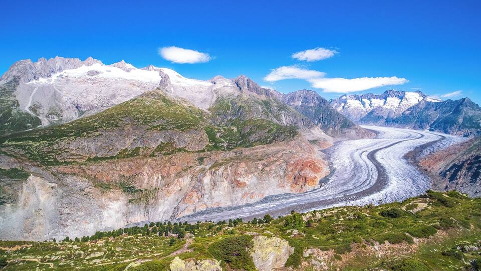 A glacier winding through a mountainous landscape with rocky outcrops and patches of greenery.