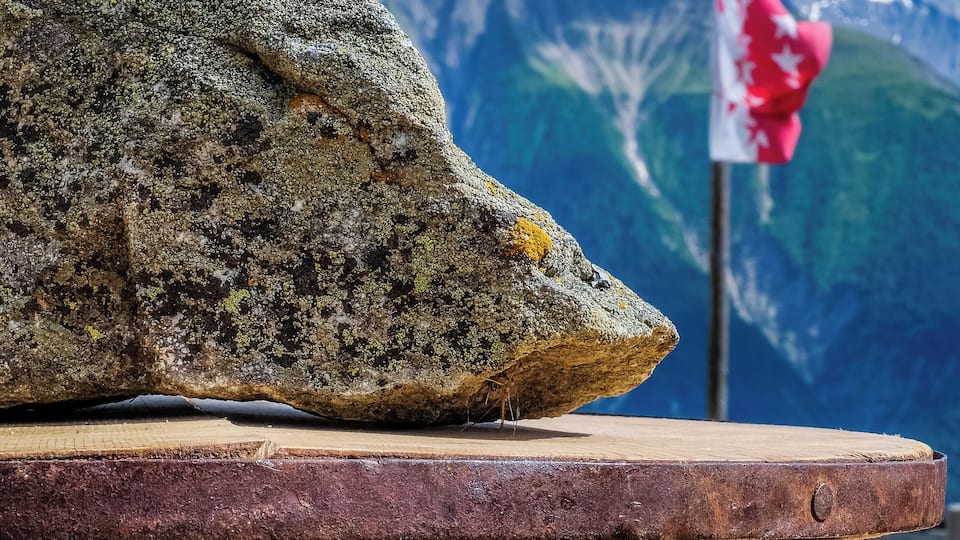 A large rock with lichen, a wooden platform, and a flag in the background.