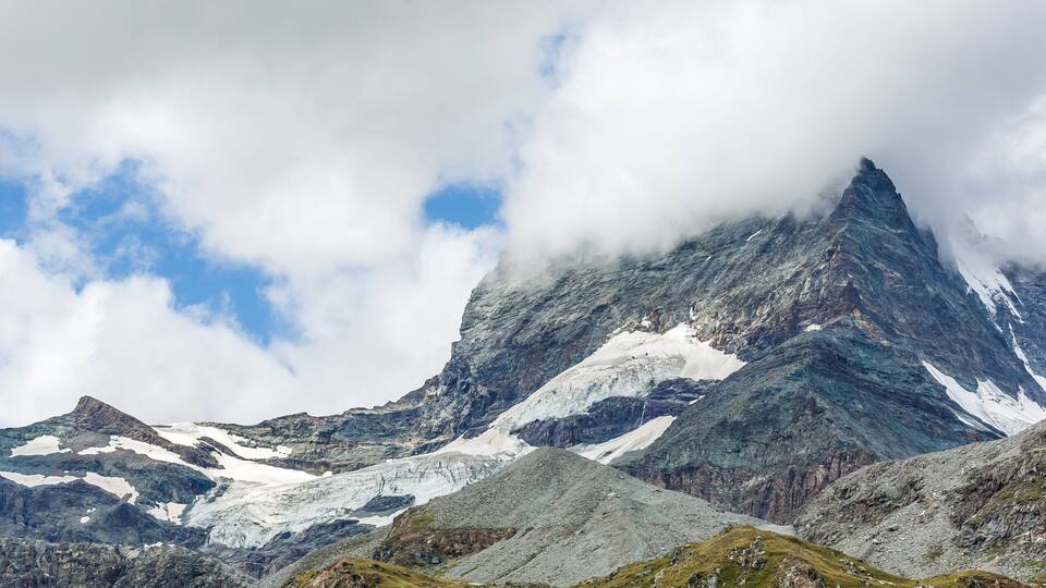 A rugged mountain range with snow patches and a prominent peak partially obscured by clouds.