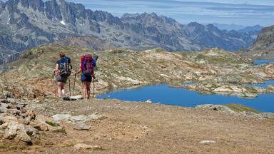 Two hikers on a rocky mountain path with a lake and distant mountains.