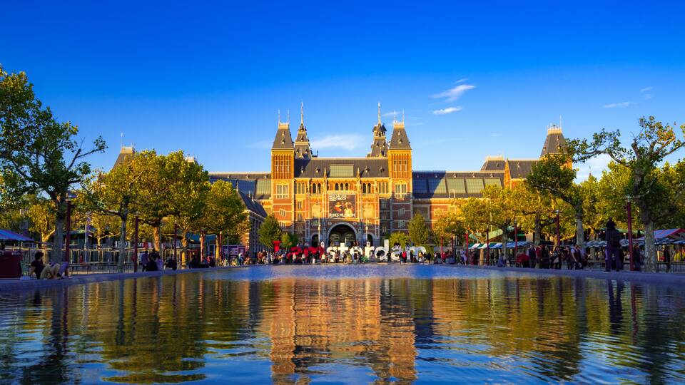 A historic building with two towers, a reflection in a body of water, and trees around the area.