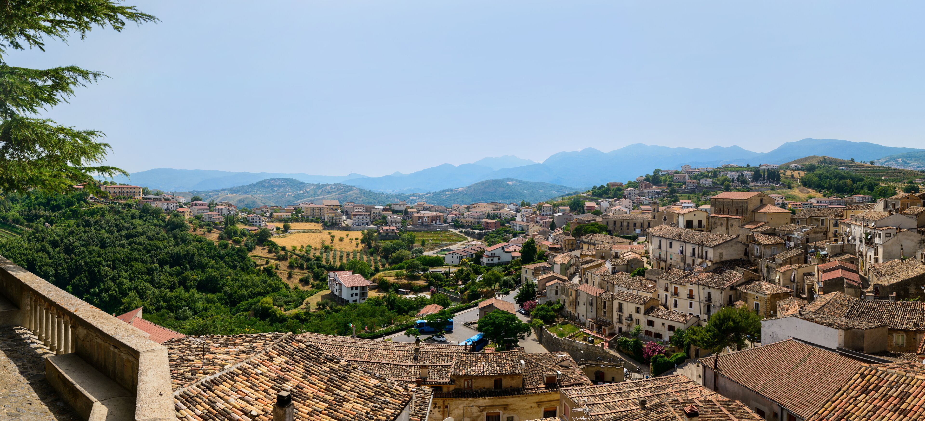 Uma cidade com edifícios de telhados em terracota, uma estrada sinuosa e montanhas ao fundo.