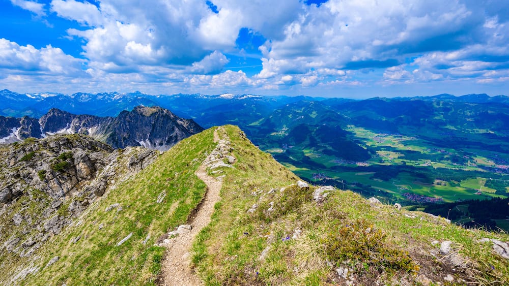 Ein Bergpfad mit Blick auf ferne, schneebedeckte Gipfel und grüne Täler.
