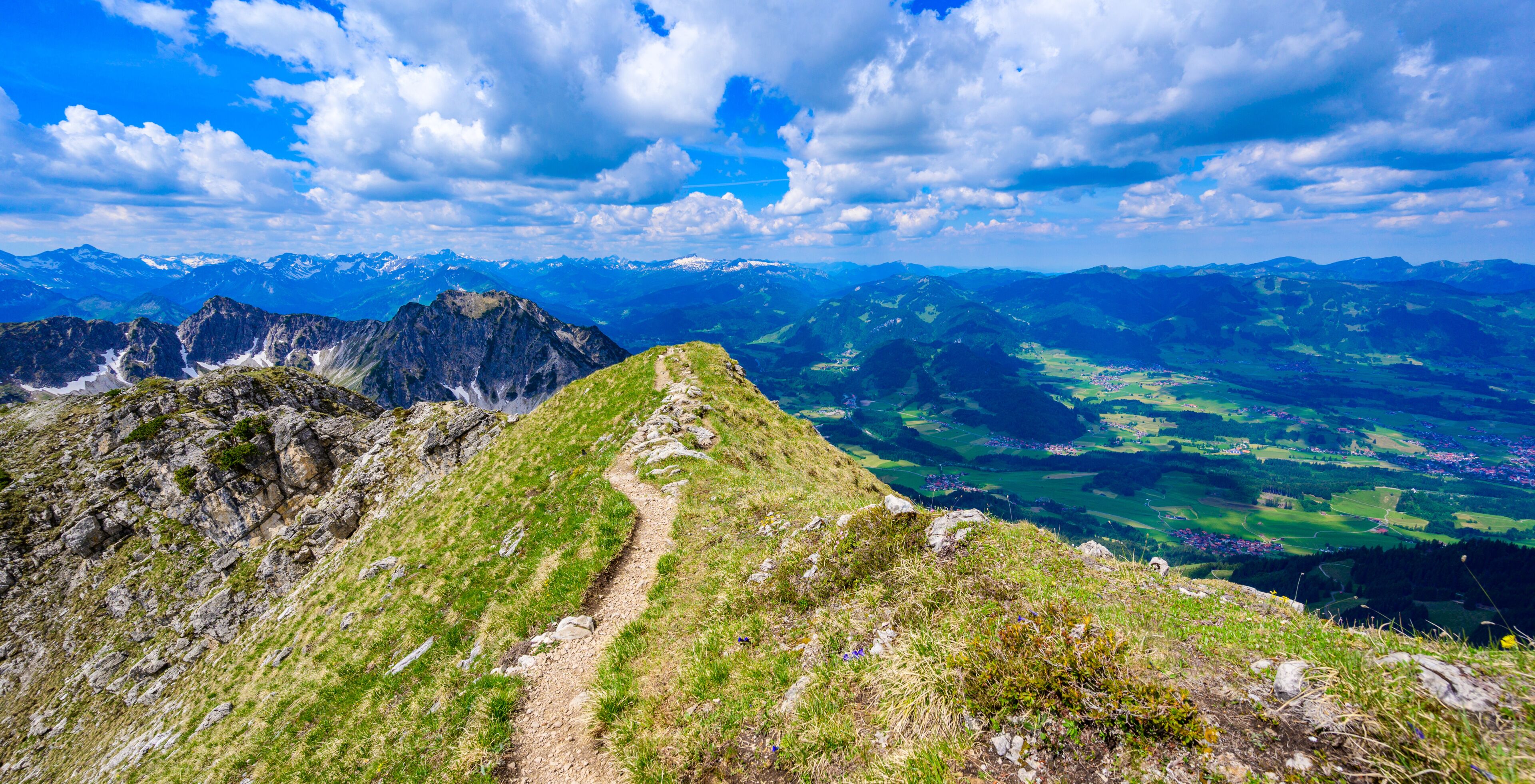 A mountain trail with a view of distant snow-capped peaks and green valleys.