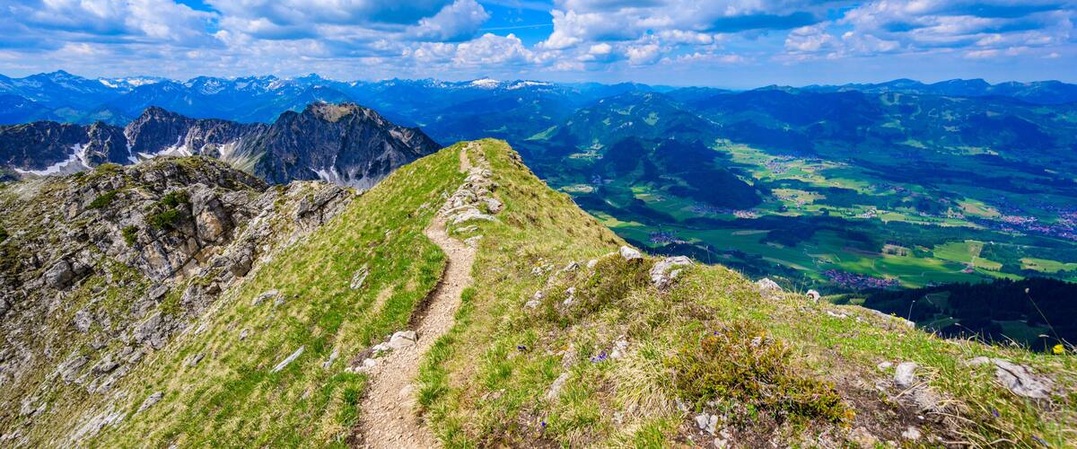 A mountain trail with a view of distant snow-capped peaks and green valleys.