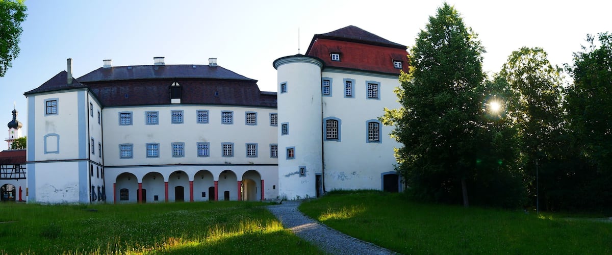 A large white building with a red roof and multiple windows, surrounded by green grass and trees.