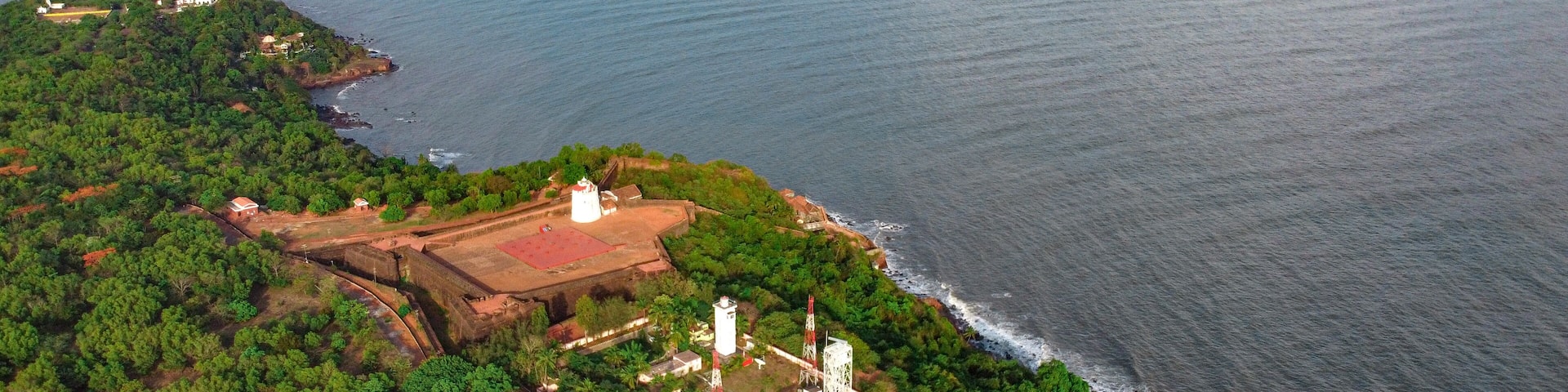 An aerial view of a coastal area with a lighthouse, dense greenery, and a calm sea.