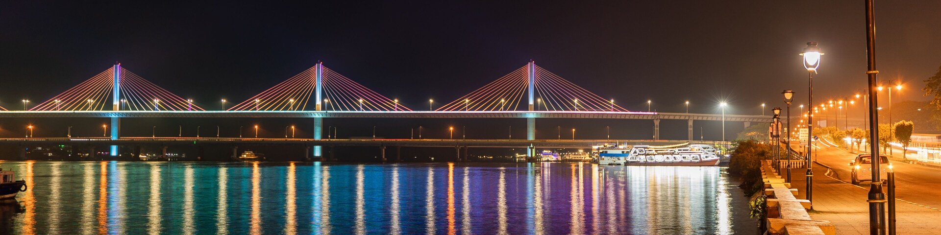 Un puente muy iluminado con varios cables, un río tranquilo que refleja las luces, y una acera con farolas.
