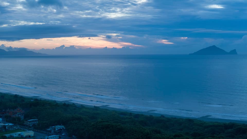 A coastal landscape with a mountain in the distance, a beach, and a small settlement with buildings.