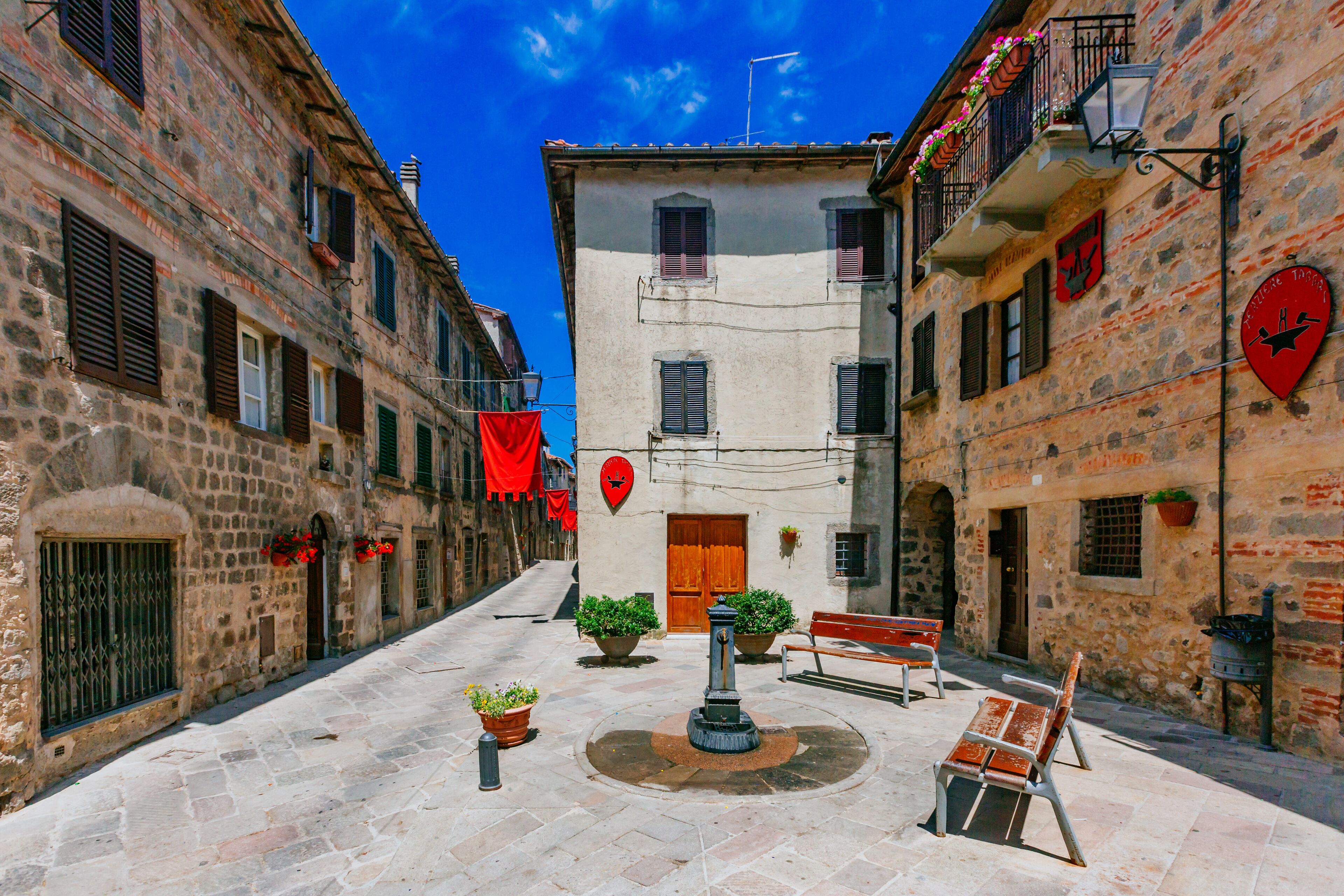 A narrow cobblestone street with stone buildings, a fountain, benches, and red decorations.