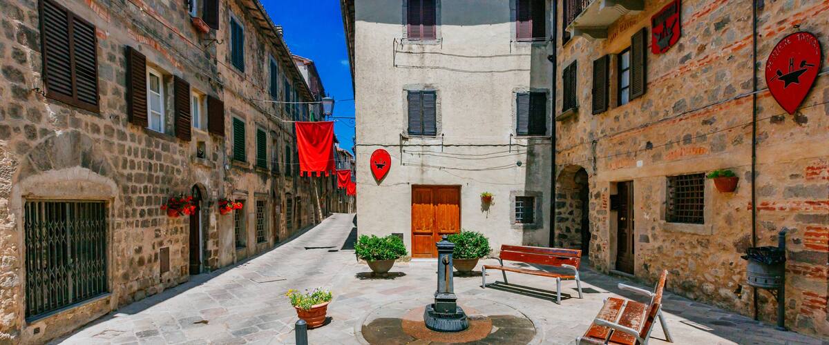 A narrow cobblestone street with stone buildings, a fountain, benches, and red decorations.