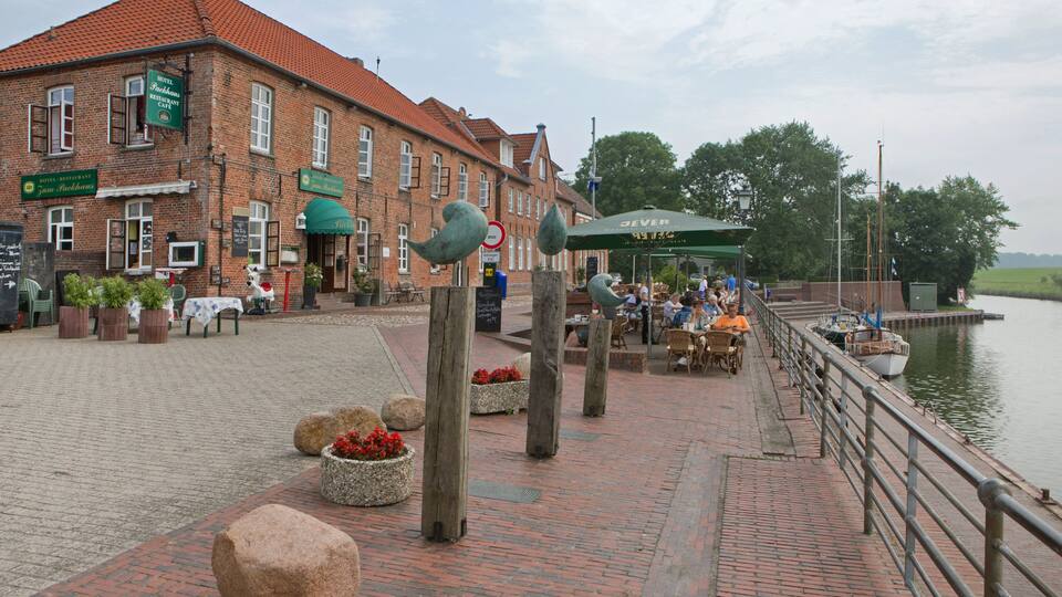 A riverside walkway with a brick path, a cafƩ, and a boat docked by the water.