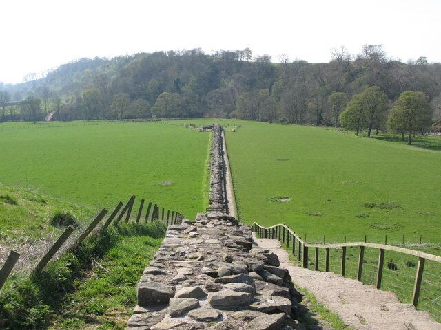 A stone pathway leading through a green field with a stone wall on one side and a fence on the other.