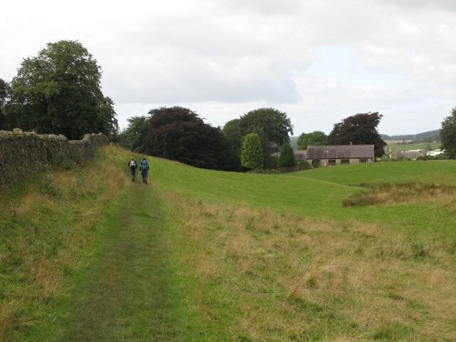 A grassy path with two hikers, a stone wall, trees, and a building in the background.