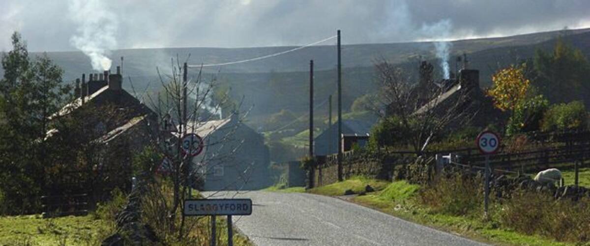 A rural road with a speed limit sign, a sign for Blagoevgrad, and smoke in the distance.