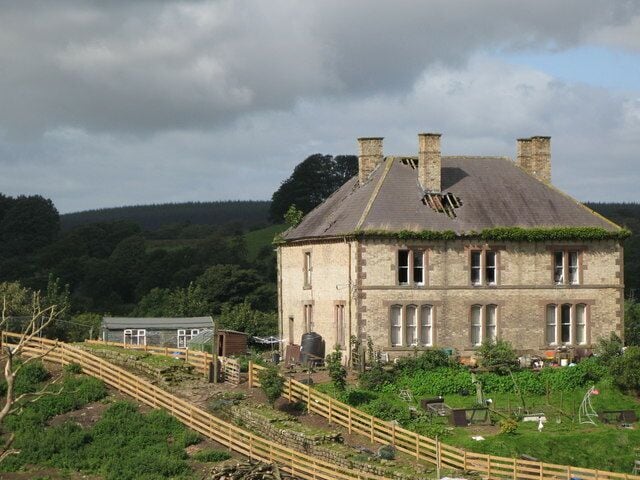 A stone house with a thatched roof, surrounded by a wooden fence and lush greenery.
