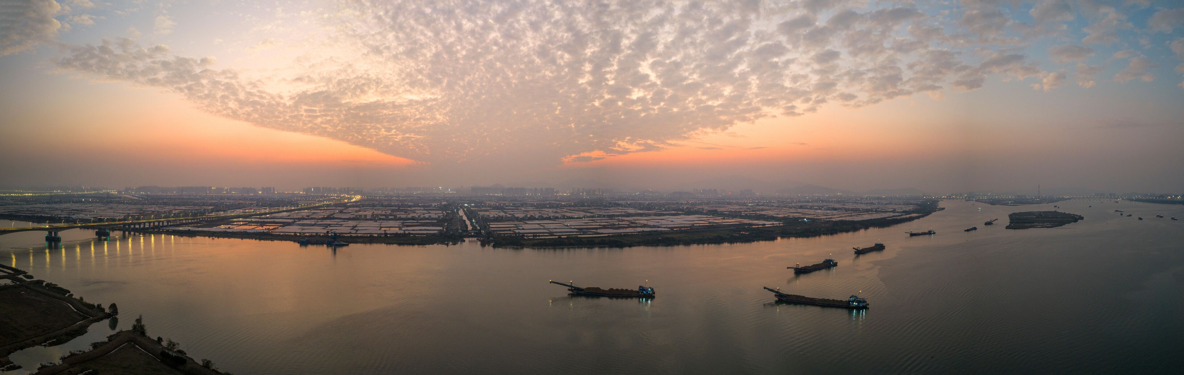A sunset over a river with boats, a bridge, and a cityscape in the distance.