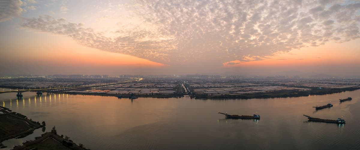 A sunset over a river with boats, a bridge, and a cityscape in the distance.