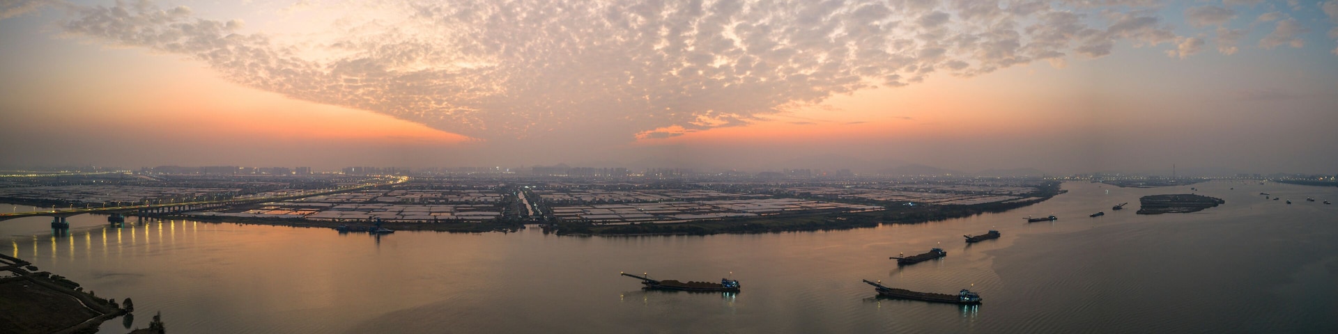 A sunset over a river with boats, a bridge, and a cityscape in the distance.