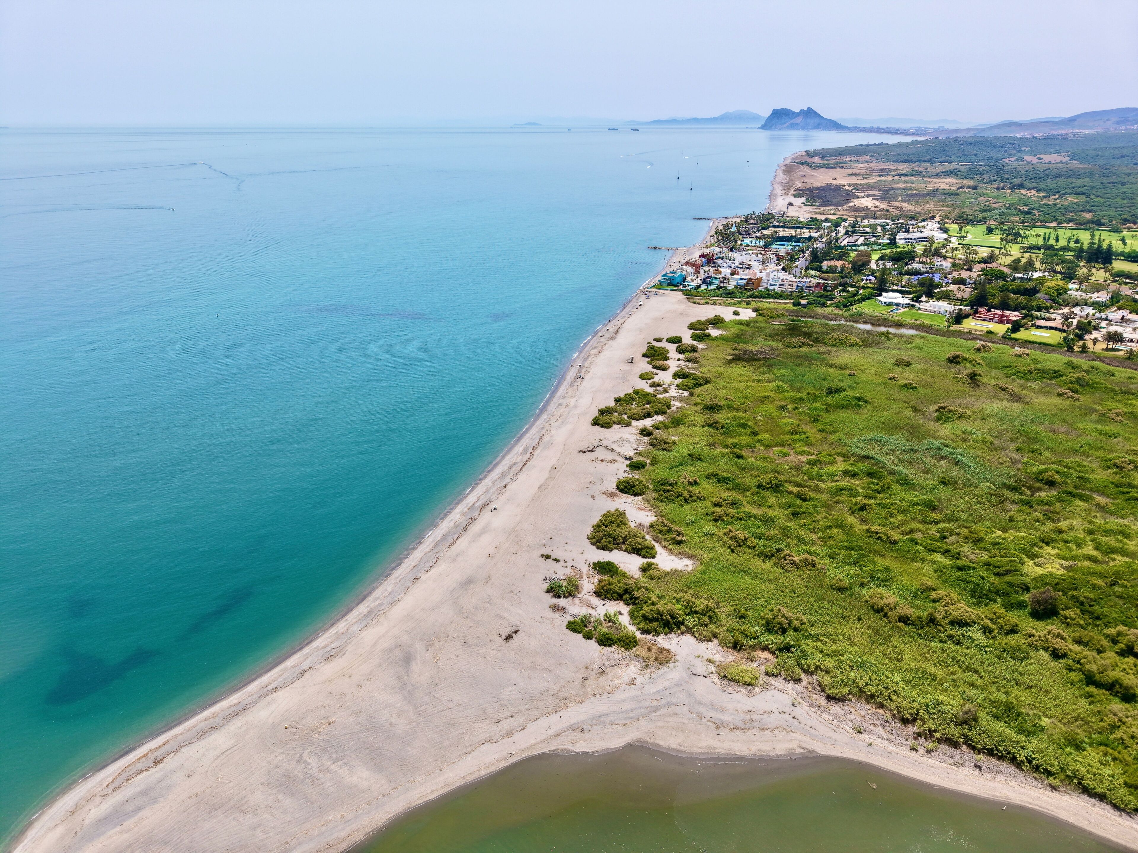 A coastal landscape with a sandy beach, a small town, and a mountain in the distance.