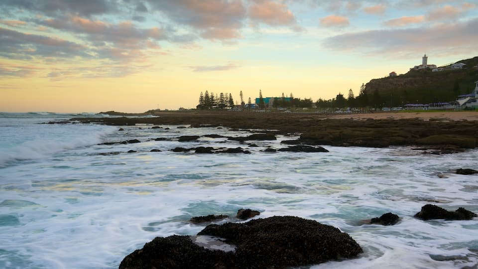A rocky shoreline with waves crashing against the rocks, a lighthouse in the distance, and a line of trees and buildings along the coast.