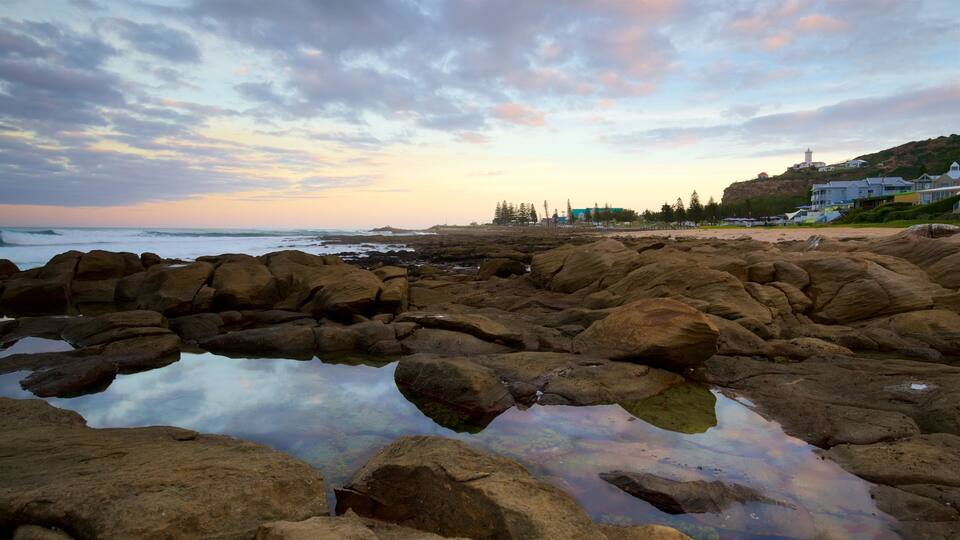 A rocky shoreline with calm water reflecting the sky, a lighthouse, and buildings on a hill in the background.