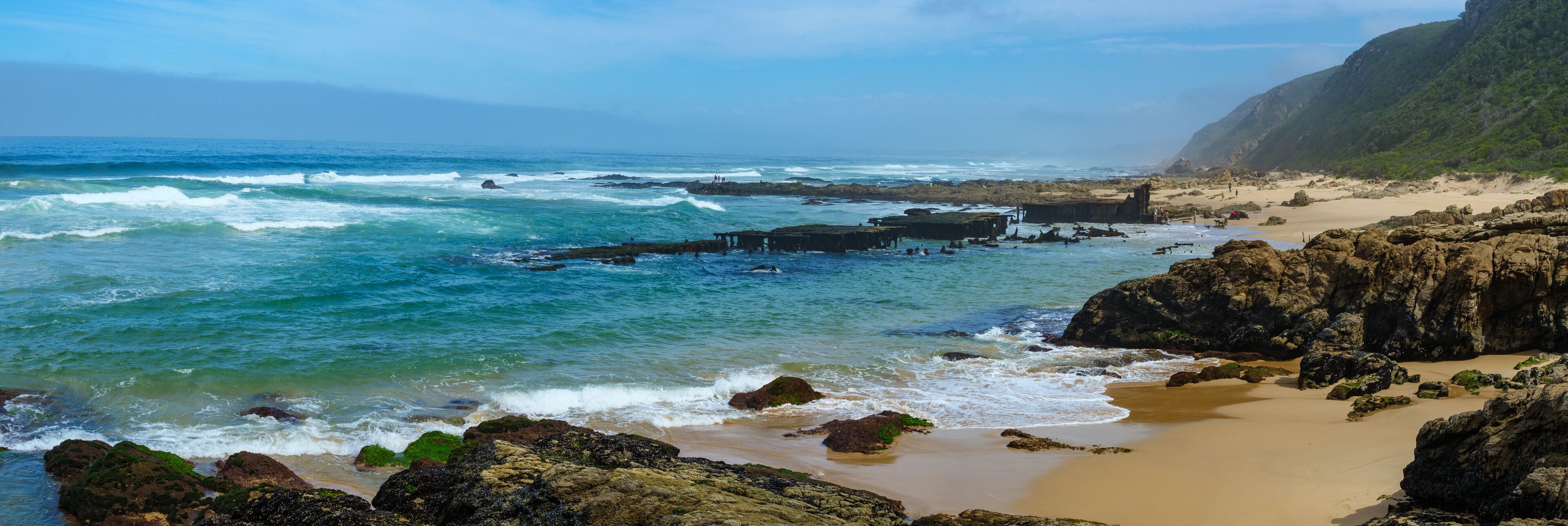 A coastal landscape with rocky shores, waves crashing onto the beach, and a clear sky.