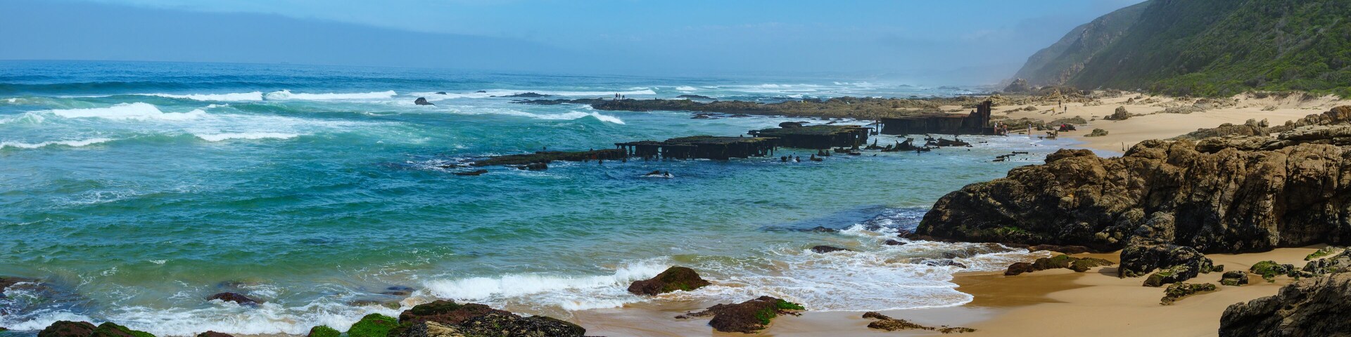 Paisagem costeira com costas rochosas, ondas quebrando na praia e céu claro.