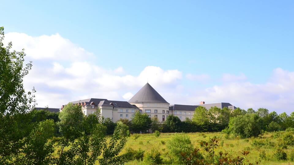 A large, multi-story building with a prominent conical roof, surrounded by lush greenery and trees.