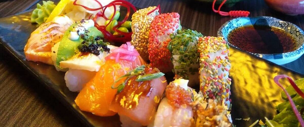 A variety of sushi on a black plate, a teapot, and a small bowl of soy sauce.