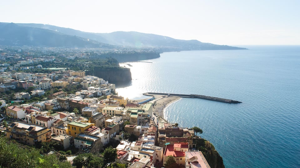 A coastal town with buildings along the shoreline, a pier extending into the sea, and mountains in the background.