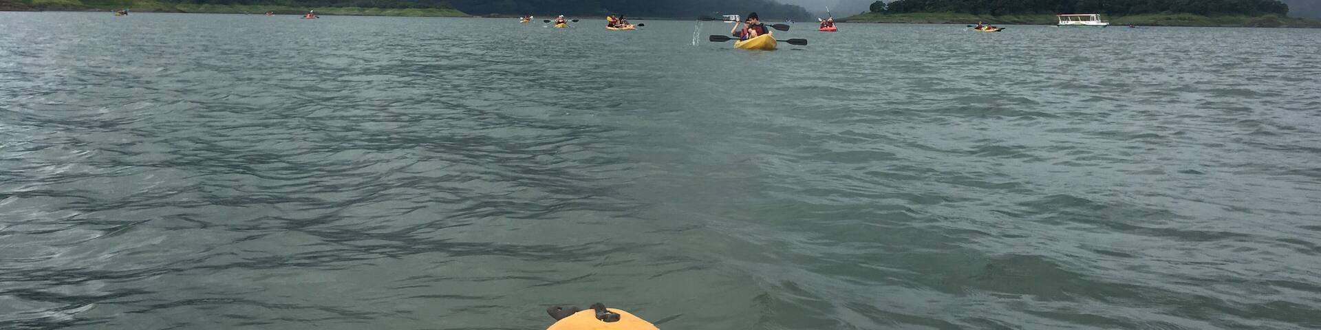 A kayak on a calm lake with other kayaks and a boat in the distance, surrounded by mountains and cloudy skies.