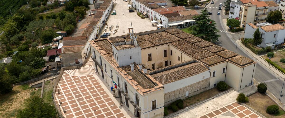 An aerial view of a historic town with a central plaza featuring a patterned pavement, surrounded by traditional buildings with terracotta roofs.