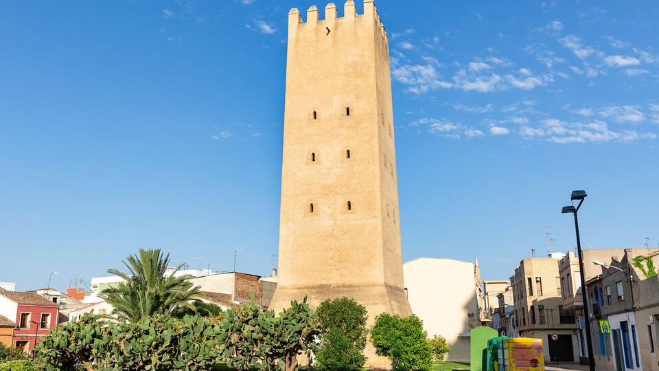 A tall, beige tower with battlements, surrounded by greenery and buildings.