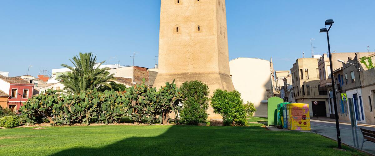 A tall, beige tower with battlements, surrounded by greenery and buildings.