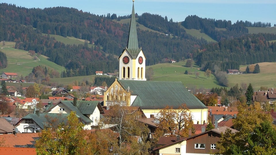 A church with a tall steeple and a clock tower, surrounded by houses and autumn-colored trees.