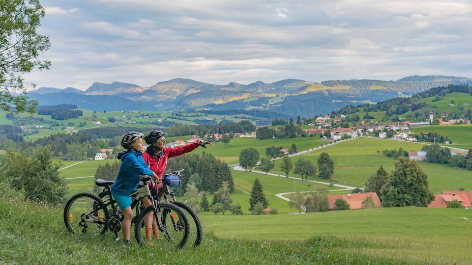 Two people on bicycles overlooking a valley with houses and mountains in the background.