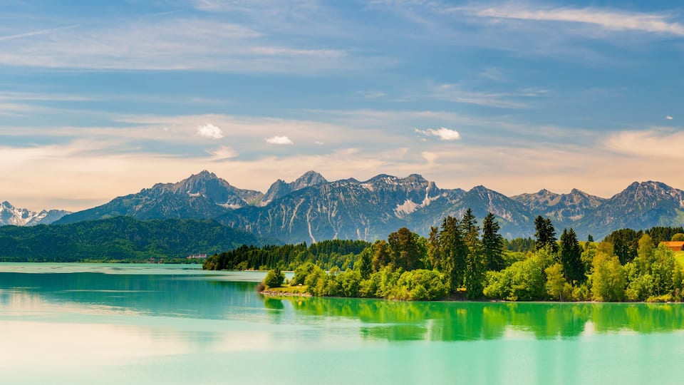 A calm lake with clear turquoise water, surrounded by lush green trees and a backdrop of snow-capped mountains under a partly cloudy sky.