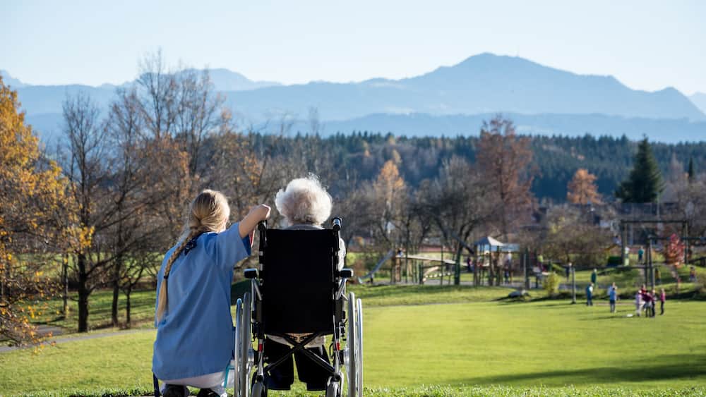 Ein Park mit einem Rollstuhl, einer Person in einem blauen Hemd und einer Bergkulisse.