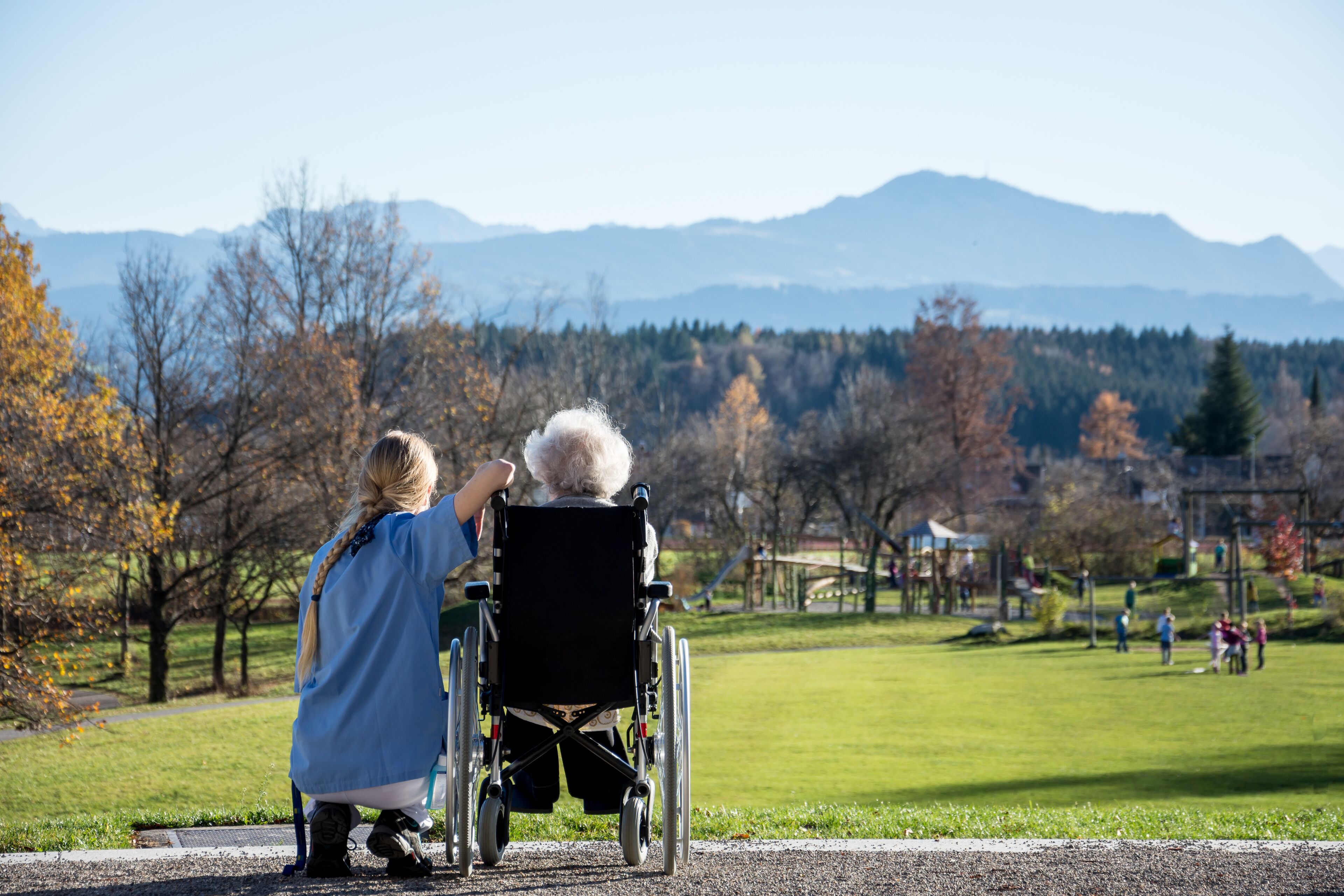 A park with a wheelchair, a person in a blue shirt, and a mountainous backdrop.