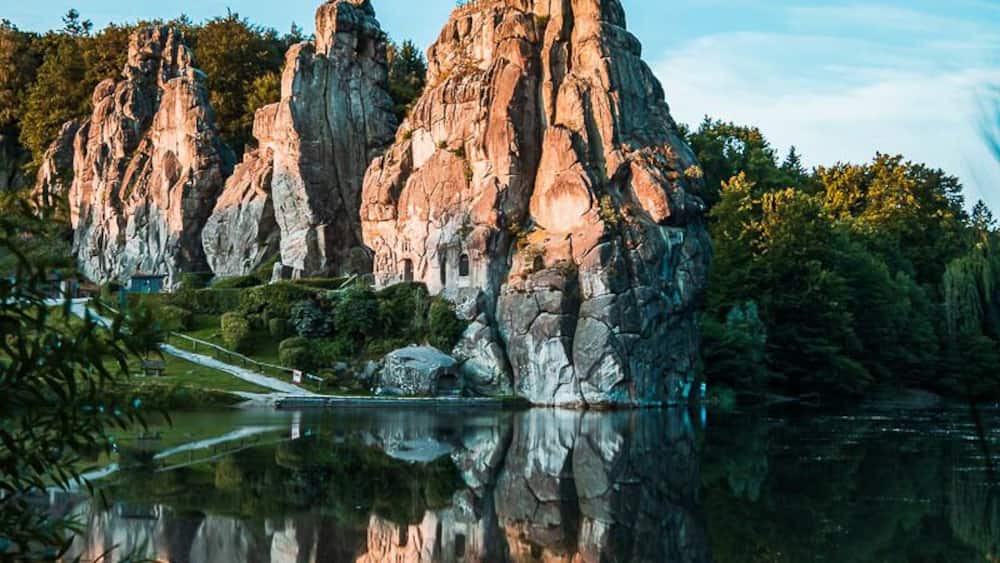 Eine felsige Insel mit deutlicher Spiegelung im ruhigen Wasser, umgeben von Grün.