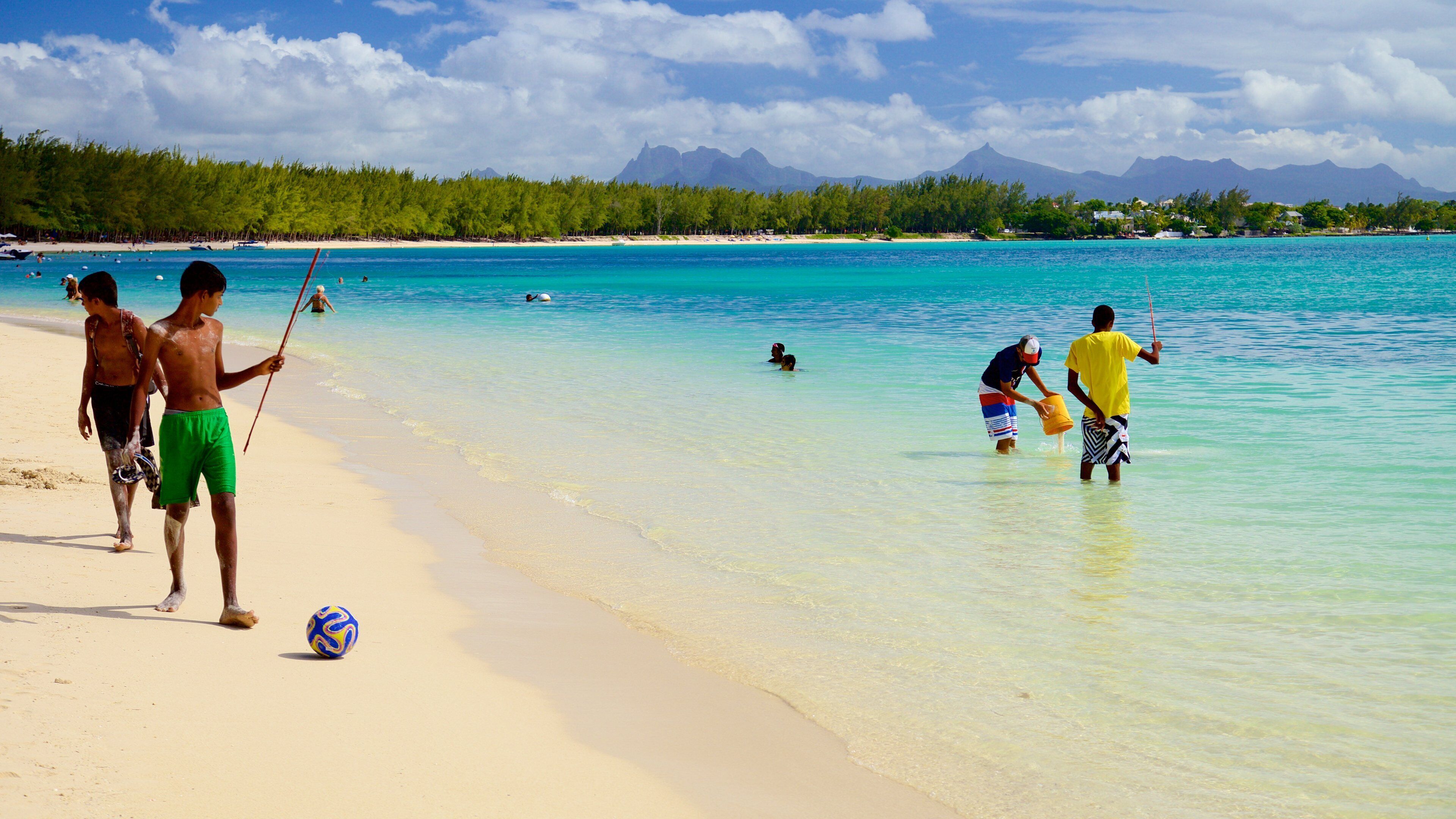 A beach with clear turquoise water, people swimming and playing, and mountains in the background.