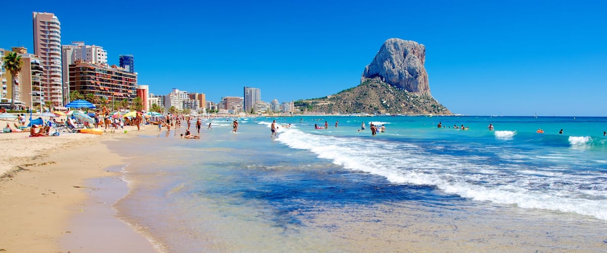 A beach with clear blue water, people swimming and sunbathing, and a prominent rocky hill in the background.