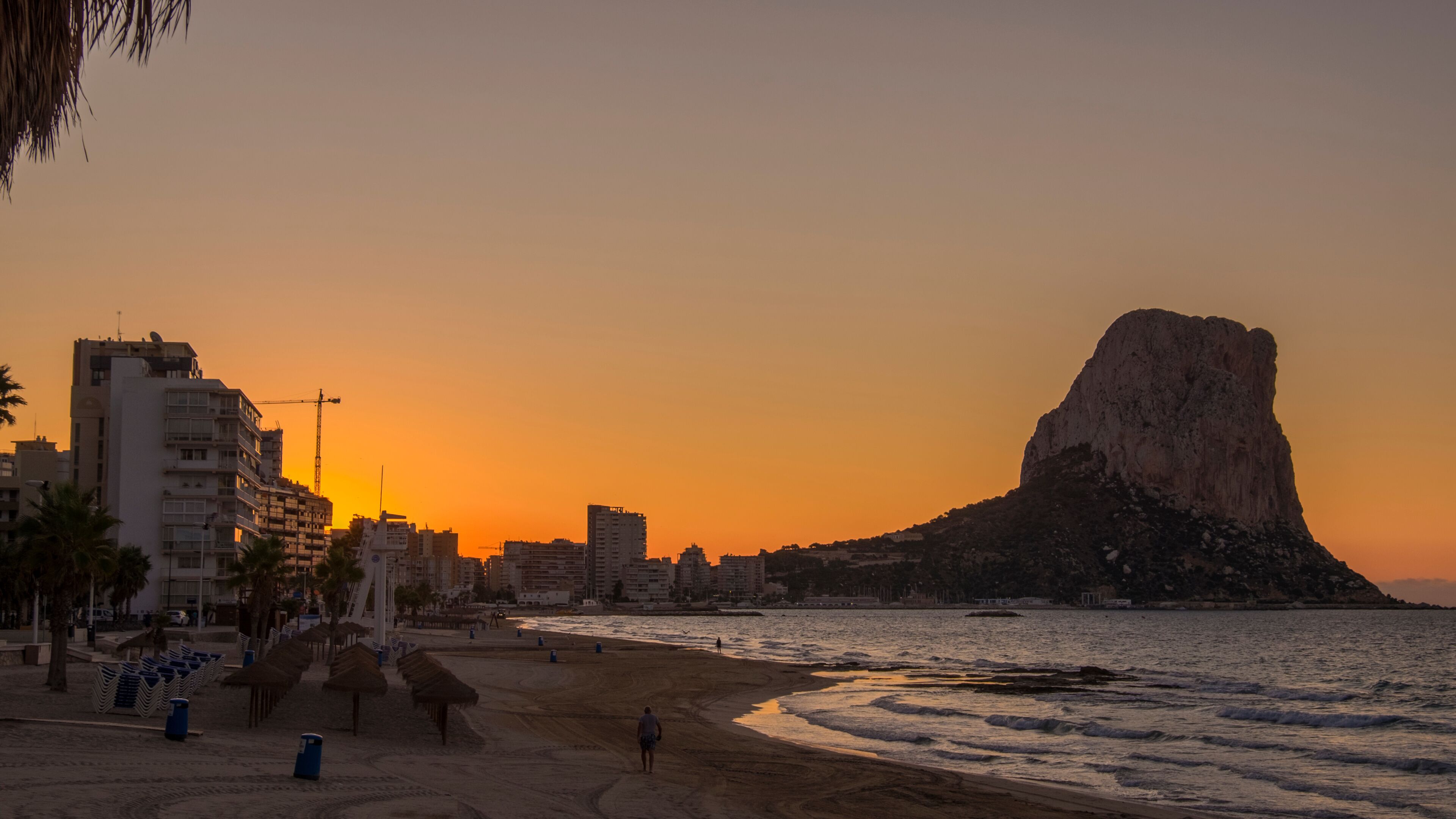 A beach with a prominent rock formation, a city skyline, and a sunset.