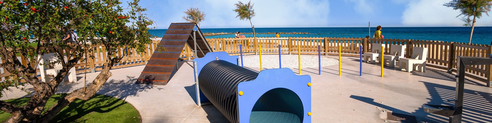 A playground with a slide, a tunnel, and a beach in the background.