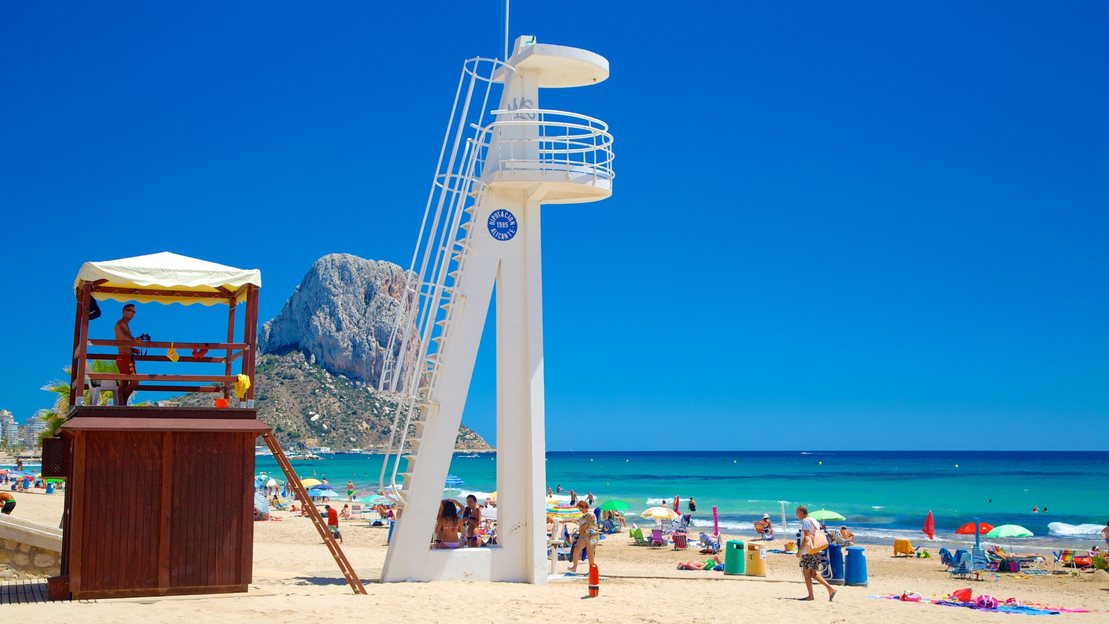 Una playa con un faro blanco alto, un puesto de socorristas de madera y gente relajándose en la arena.