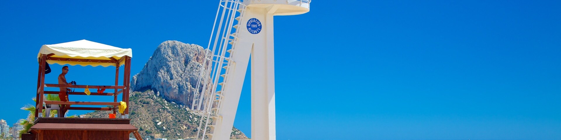 Una playa con un faro blanco alto, un puesto de socorristas de madera y gente relajándose en la arena.